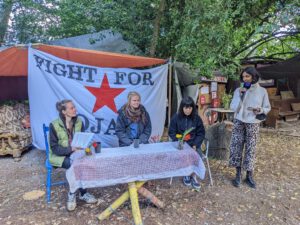 Drei weiblich gelesene Menschen vor einem Transpi mit der Aufschrift "Fight for Rojava" , eine weiblich gelesene Person mit Mikrofon
